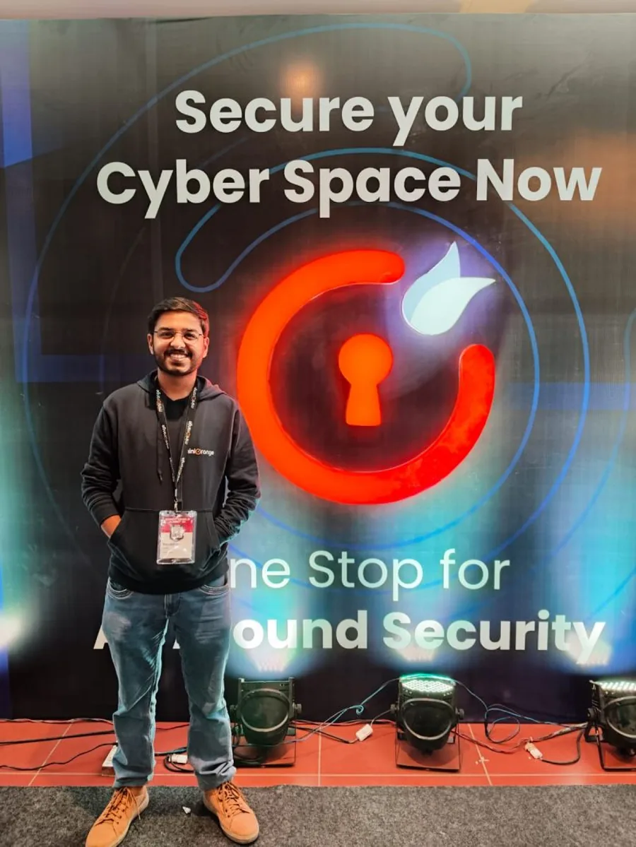 A man in a black hoodie smiling in front of the Secure your Cyber Space Now panel with the lit miniOrange logo